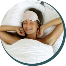 A woman with a sleep mask on her forehead, waking up stretching and looking happy.