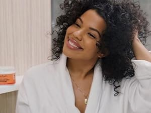 Woman with curly hair, smiling with hand in hair, SheaMoisture smoothie container in background.