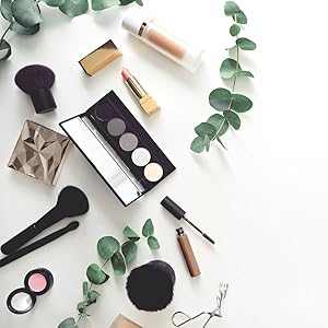 Flatlay of makeup and beauty accessories including brushes, eyeshadow palette, and eucalyptus sprigs on white background